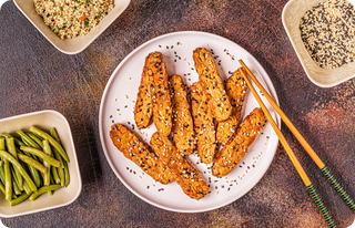 A plate of tempeh slices topped with sesame seeds, surrounded by bowls of snap peas, cooked grains, and more sesame seeds. Chopsticks rest beside the plate on a textured brown surface.
