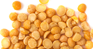 Close-up of yellow split peas scattered on a white background. Some peas are whole and round, while others are broken and flat, displaying their smooth surface and distinct yellow color.
