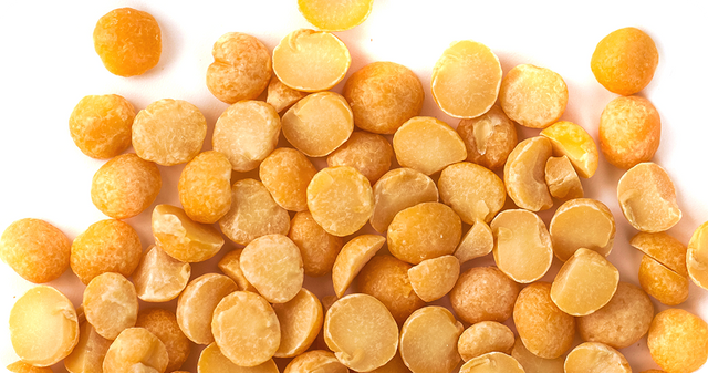 Close-up of yellow split peas scattered on a white background. Some peas are whole and round, while others are broken and flat, displaying their smooth surface and distinct yellow color.