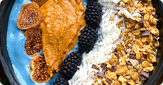 A blue bowl contains sliced figs, blackberry clusters, shredded coconut, granola with chocolate chunks, and a dollop of nut butter, arranged in neat rows.