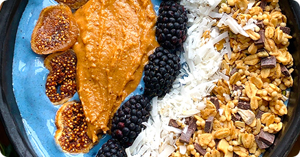 A blue bowl contains sliced figs, blackberry clusters, shredded coconut, granola with chocolate chunks, and a dollop of nut butter, arranged in neat rows.