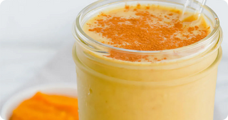A glass of creamy orange smoothie topped with a sprinkle of cinnamon, with a clear straw and a blurred orange ingredient in the background.