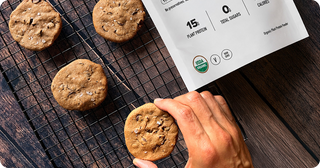 A hand picks up a chocolate chip cookie from a cooling rack with four cookies, beside a nutrition label showing 15g plant protein, 0g sugar, and other information. The scene is set on a dark wooden surface.