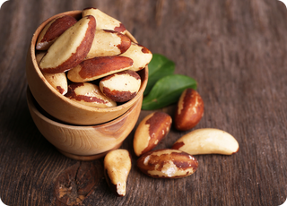 A wooden bowl filled with Brazil nuts sits on a rustic wooden surface, with a few nuts scattered around and green leaves placed nearby.