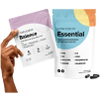 A hand holds a purple and white supplement packet labeled Essential. Next to it is a larger blue and white Essential pouch with vitamins and capsules displayed nearby.