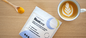 A packet labeled Complement Restore, a spoonful of yellow powder, and a cup of latte with leaf-shaped foam art are arranged on a wooden table.
