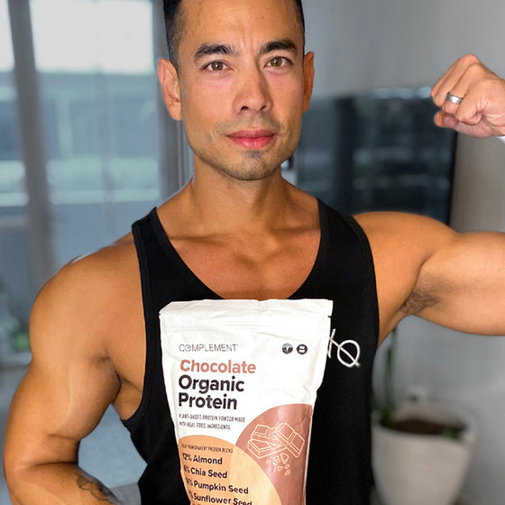 A person flexes their bicep while holding a bag of chocolate organic protein powder. They are wearing a black tank top, standing indoors with blurred background elements like furniture and plants.
