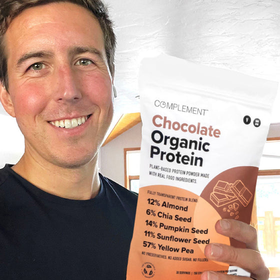 A person is smiling and holding a bag of Complement Chocolate Organic Protein powder. The packaging lists ingredients: almond, chia seed, pumpkin seed, sunflower seed, and yellow pea. The background features a bright interior with wooden accents.