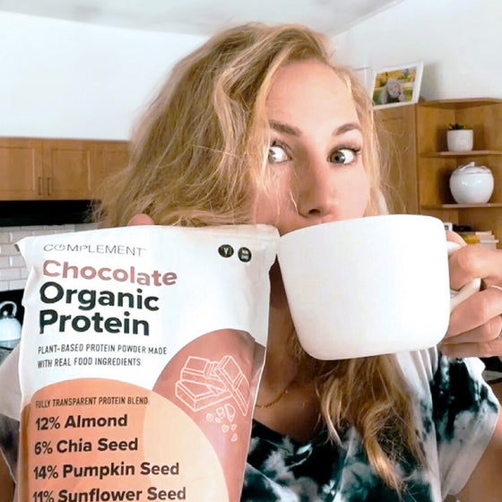 A person with long blonde hair holds a large white mug in one hand and a bag of chocolate organic protein powder in the other. They appear to be in a kitchen with wooden cabinets in the background.
