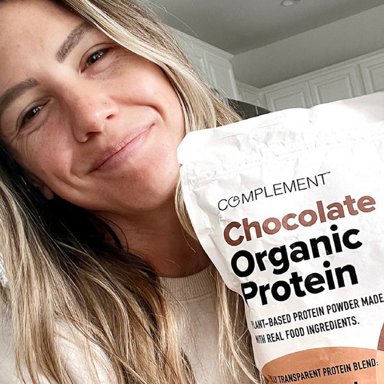A person smiling and holding a bag of Complement Chocolate Organic Protein powder. The background shows a kitchen.