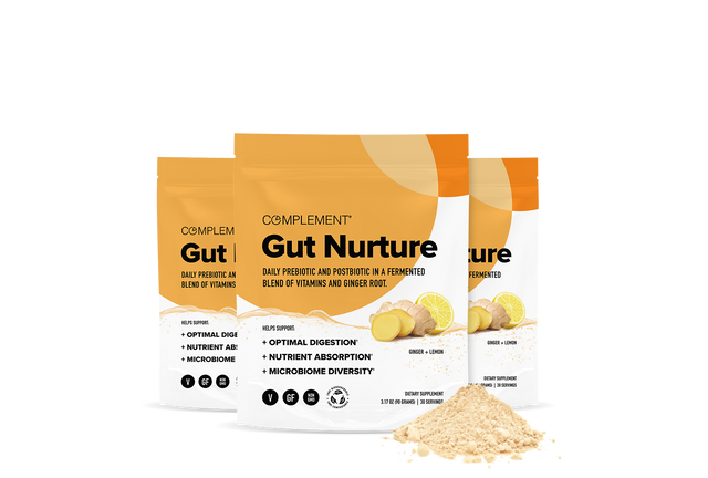 Three orange and white bags labeled Complement Gut Nurture with images of ginger and powder in front, promoting gut health benefits. A small mound of beige powder is displayed near the bags.