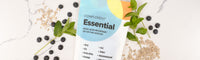 A Complement Essential multivitamin pouch lies on a marble surface surrounded by blueberries, sunflower seeds, brown rice, and mint leaves. The product label highlights key nutrients for a plant-based diet.