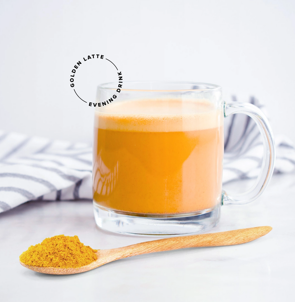 A glass mug of golden latte on a white surface with a striped cloth in the background. A wooden spoon with turmeric powder is in the foreground. Text reads Golden Latte Evening Drink.