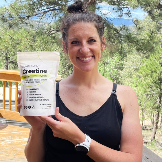 A smiling woman in a black tank top stands outdoors, holding a bag of Complement Creatine supplement. Trees and mountains are visible in the background.