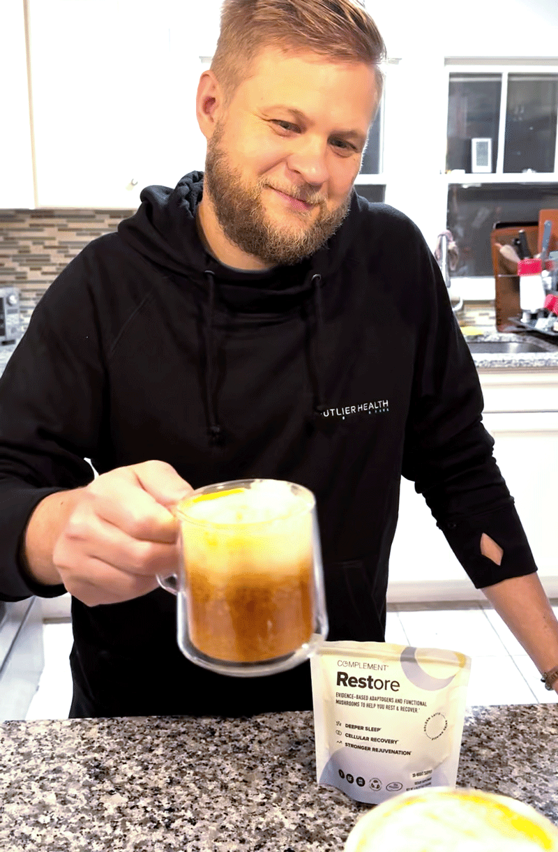 A smiling man with a light beard holds a glass mug of frothy brown drink in a kitchen. On the counter in front of him is a packet labeled Restore. Kitchen items are visible in the background.
