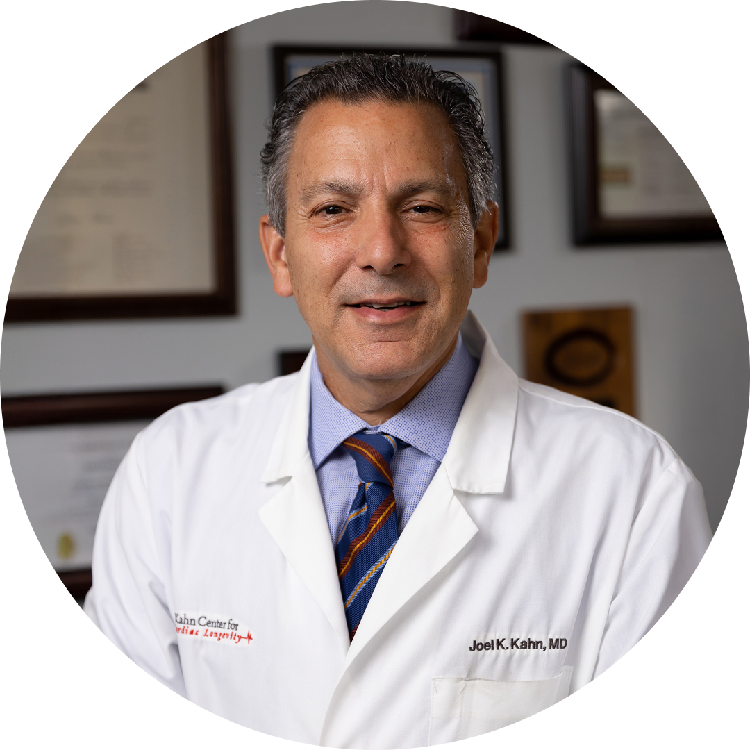A middle-aged man in a white lab coat with Joel K. Kahn, MD embroidered, smiling and standing in an office with framed certificates and awards on the wall behind him.