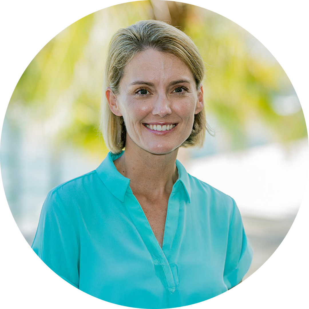 A woman with short blonde hair wearing a teal blouse smiles at the camera outdoors, with blurred greenery and sunlight in the background.