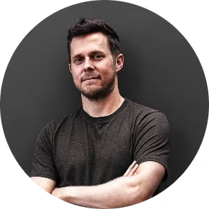 A man with short dark hair and facial stubble stands against a dark background. He is wearing a dark t-shirt and has his arms crossed, giving a relaxed yet confident expression.