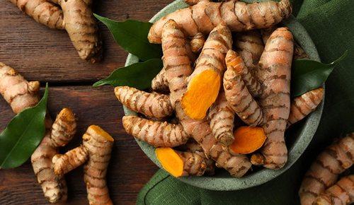 A bowl of fresh turmeric roots sits on a wooden table. Some roots are sliced to reveal their bright orange interior. Green leaves accent the arrangement, and a green cloth partially covers the table surface.