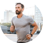 A man with a beard jogging outdoors in an urban setting, wearing a gray t-shirt and a smartwatch. Buildings and trees are blurred in the background, suggesting motion.