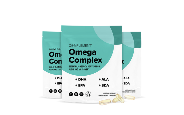 Three teal and white pouches labeled Complement Omega Complex with supplement information are displayed upright, with three transparent capsules placed in front of them on a white background.