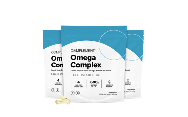 Three blue and white Complement Omega Complex supplement bags stand upright with two yellow capsules in front; the packaging highlights vegan omega-3, 600mg DHA+EPA, and key features like being sourced from algae.
