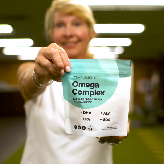 A smiling woman in a white shirt holds up a package of Complement Omega Complex supplement, with the branding and nutrient information visible on the front of the pouch. The background is softly blurred.