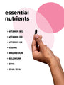 Hand holding a black capsule against a white background with pink abstract shapes. Text listing essential nutrients: Vitamin B12, D3, K2, Iodine, Magnesium, Selenium, Zinc, DHA/EPA.