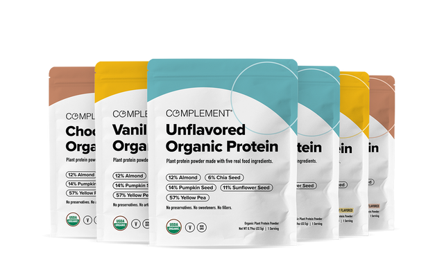 Five colorful pouches of Complement Organic Protein powder are displayed in a row, with flavors including Unflavored, Vanilla, and Chocolate. Each pouch lists main ingredients like almond, pumpkin seed, sunflower seed, and yellow pea.