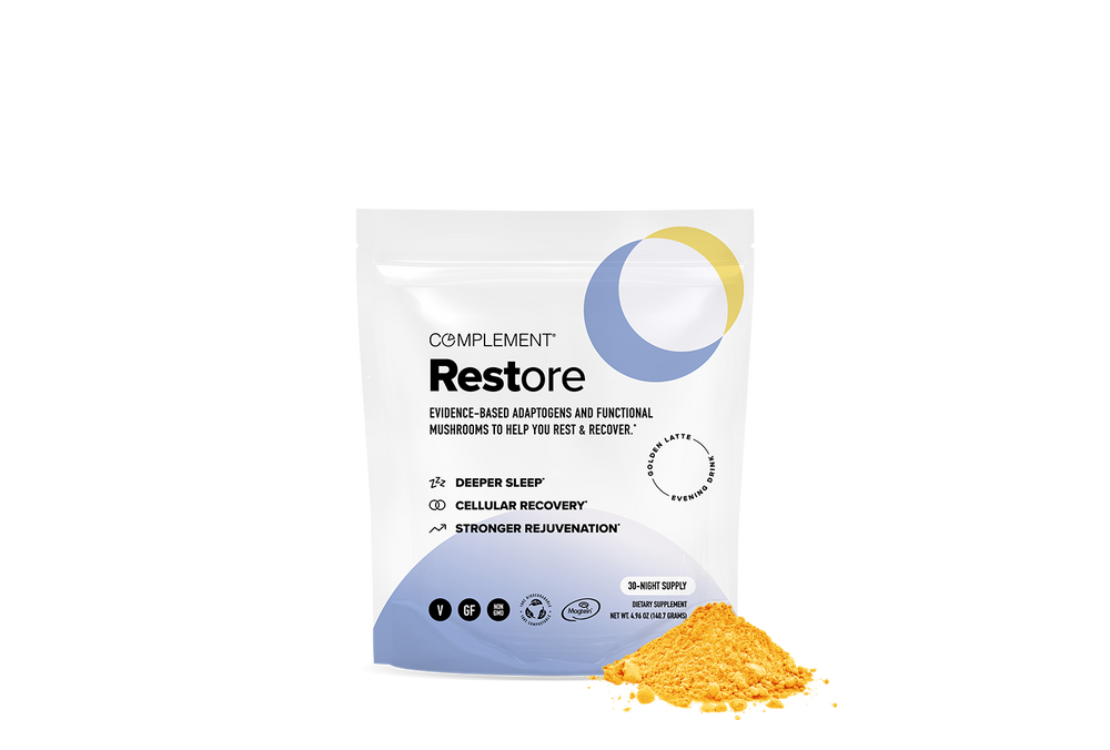 A white pouch labeled Complement Restore with text highlighting benefits like deeper sleep and cellular recovery, sits next to a small mound of yellow-orange powder. The packaging features blue and yellow circle designs.