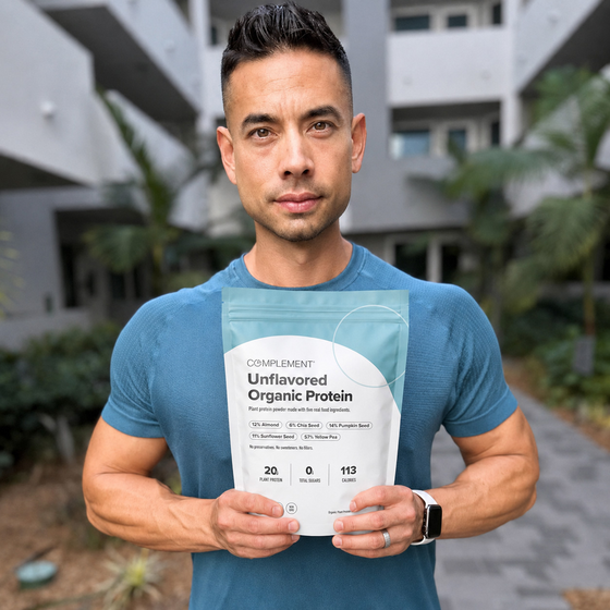 A man in a blue t-shirt stands outdoors, holding a bag of Complement Unflavored Organic Protein. An apartment building and palm trees are visible in the background.