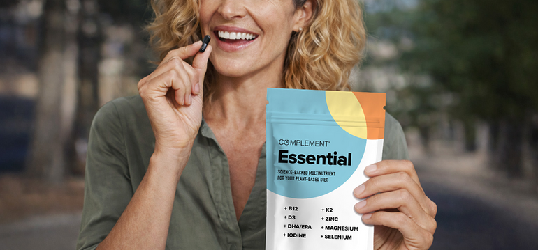 A smiling woman holds a supplement capsule in one hand and a bag labeled Complement Essential with various nutrients listed in the other hand, outdoors.