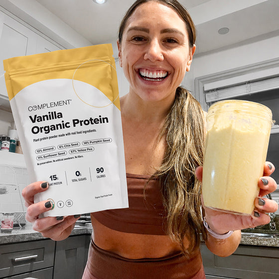 A smiling woman stands in a kitchen holding a large protein shake and a bag of vanilla organic protein powder, showing the front label.