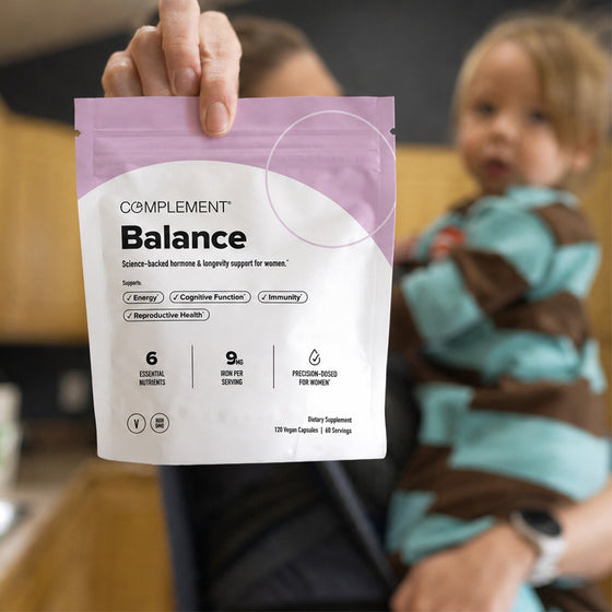A person holds a bag labeled Complement Balance dietary supplement, designed for women, in the foreground. In the background, another person holds a young child in a kitchen setting.