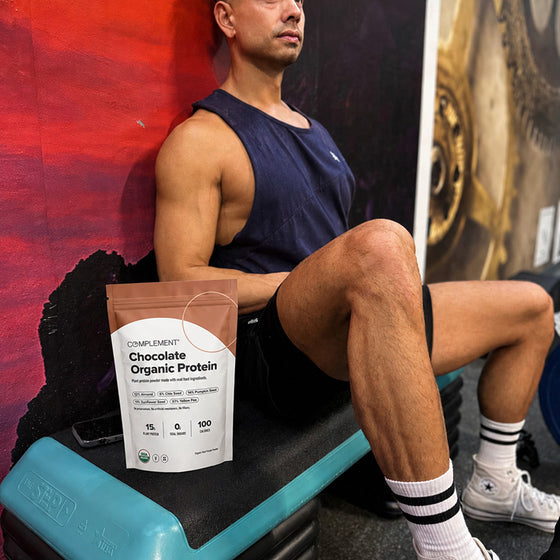 A man in workout clothes sits on a gym bench next to a bag of Complement Chocolate Organic Protein, with gym equipment and a colorful wall in the background.