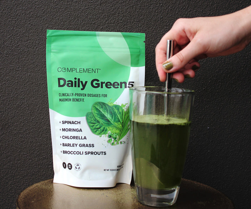 A hand stirs a glass of green smoothie with a metal straw next to a green and white bag labeled Complement Daily Greens featuring leafy greens and listing ingredients like spinach, moringa, and chlorella.