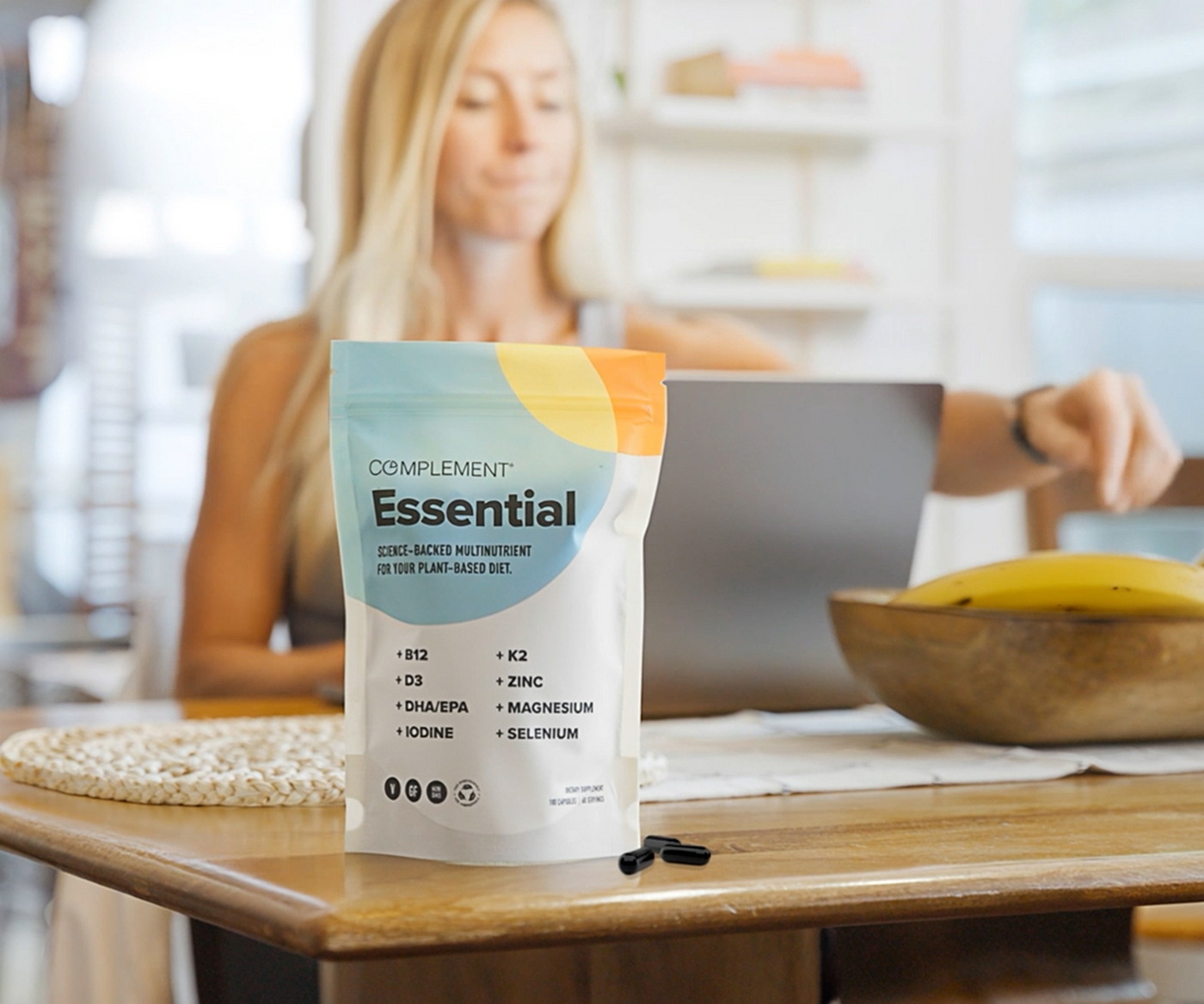 A pouch of Complement Essential plant-based nutrients stands on a wooden table with capsules beside it; a woman works on a laptop in the blurred background with a bowl of bananas nearby.