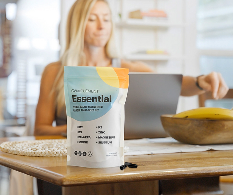 A pouch of Complement Essential plant-based nutrients stands on a wooden table with capsules beside it; a woman works on a laptop in the blurred background with a bowl of bananas nearby.