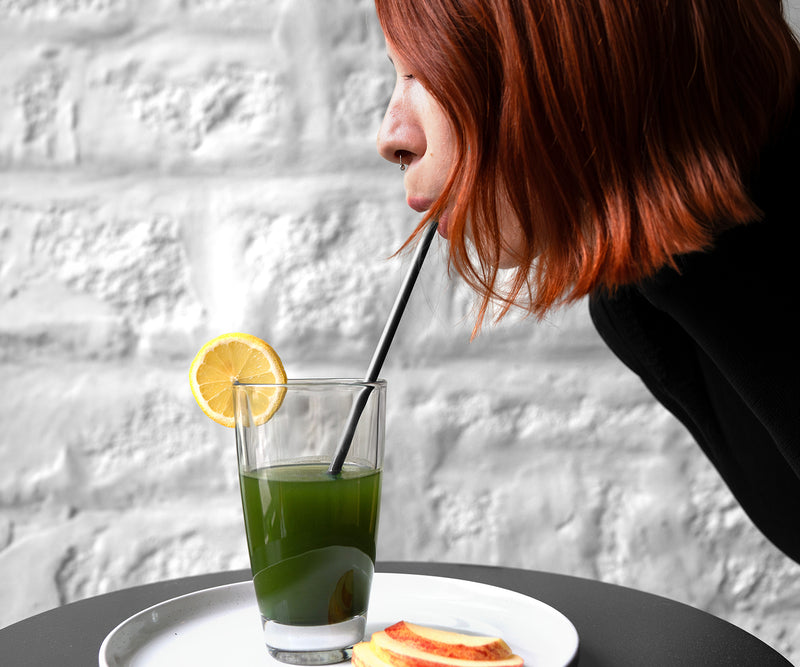 A person with red hair drinks a green beverage with a lemon slice in a glass using a straw. The glass sits on a white plate with lemon slices, against a white brick wall background.