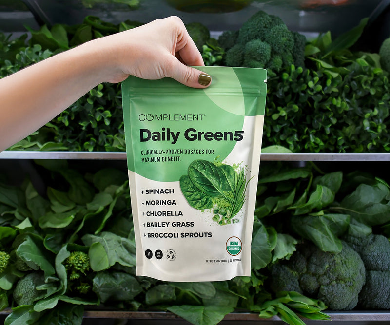 A hand holds a bag of Complement Daily Green 5 supplement in front of a display of fresh green vegetables, including broccoli, spinach, and leafy greens. The package highlights its plant-based ingredients.