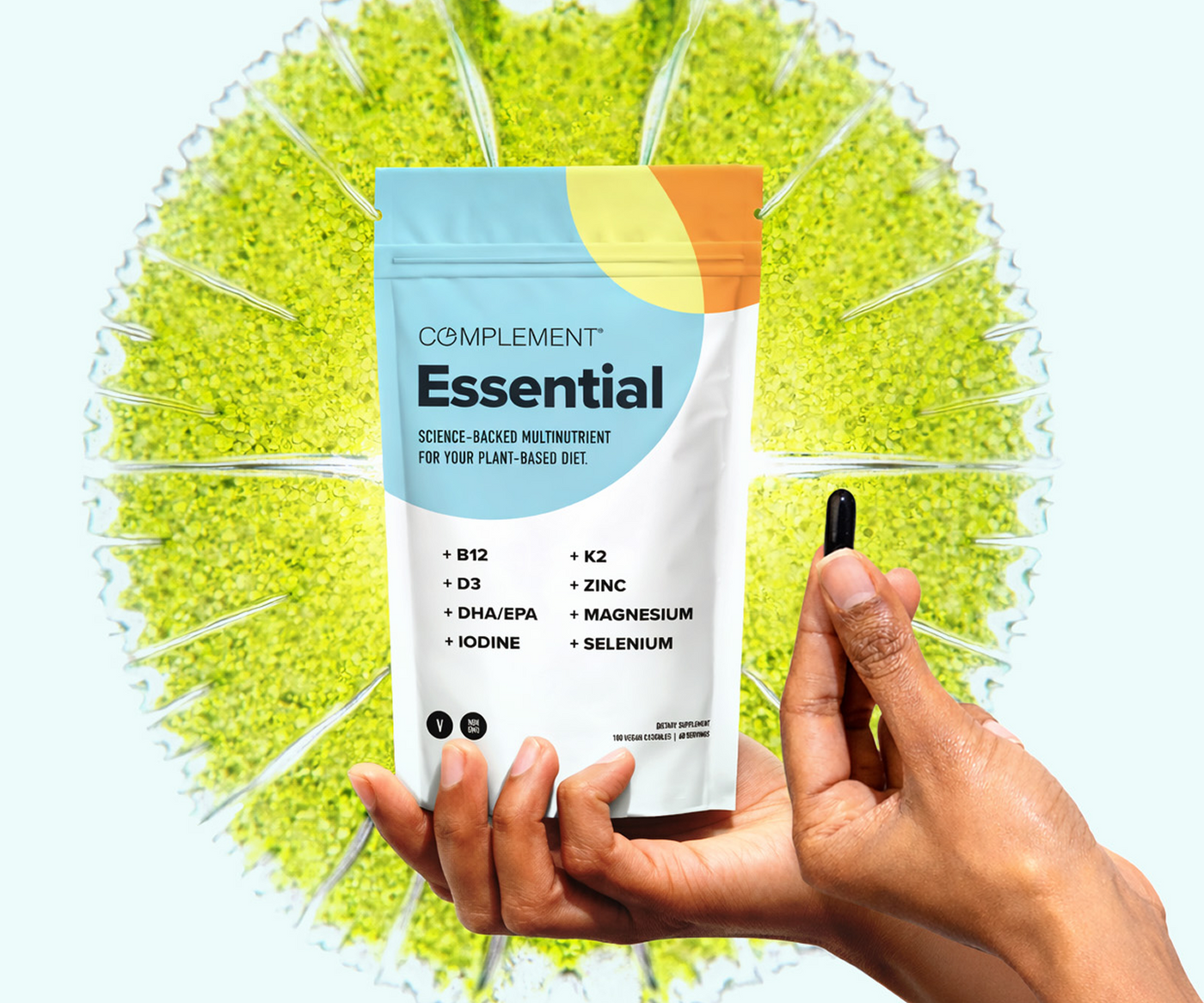 A hand holds a black capsule next to a colorful pouch labeled Complement Essential with listed nutrients, set against a bright green circular background resembling a plant or algae.