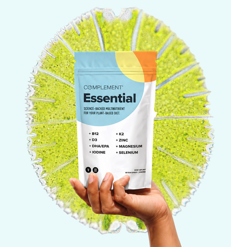 A hand holds a bag of Complement Essential multivitamins against a bright, abstract background featuring a green circular pattern.