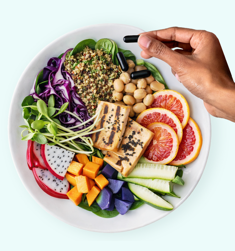 A hand holds black capsules above a plate of colorful food, including tofu, chickpeas, quinoa, spinach, purple cabbage, dragon fruit, orange slices, zucchini, purple potatoes, and cubed orange vegetables.