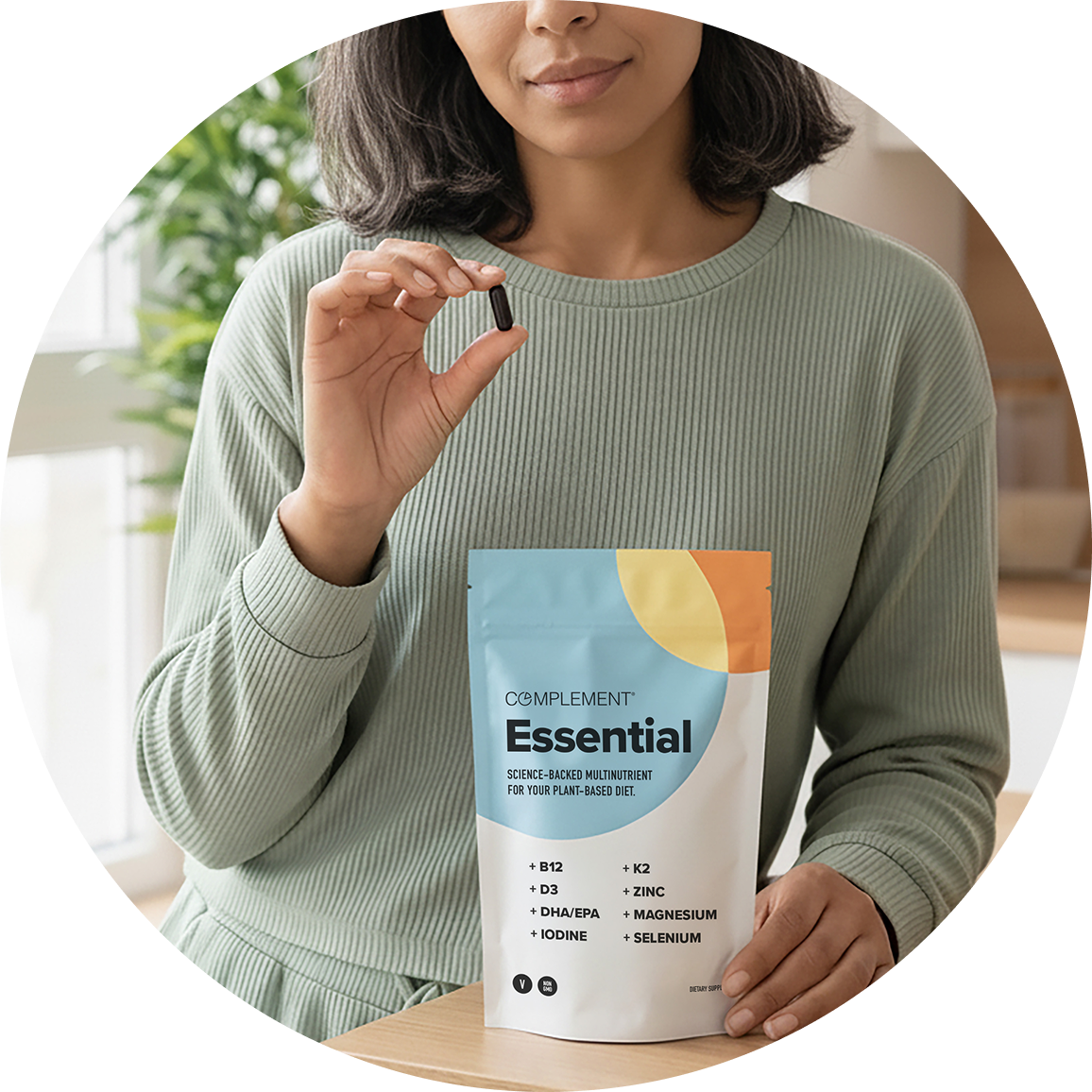 A woman wearing a light green sweater holds a capsule in one hand and a packet labeled Complement Essential multinutrient in the other, sitting indoors at a table.