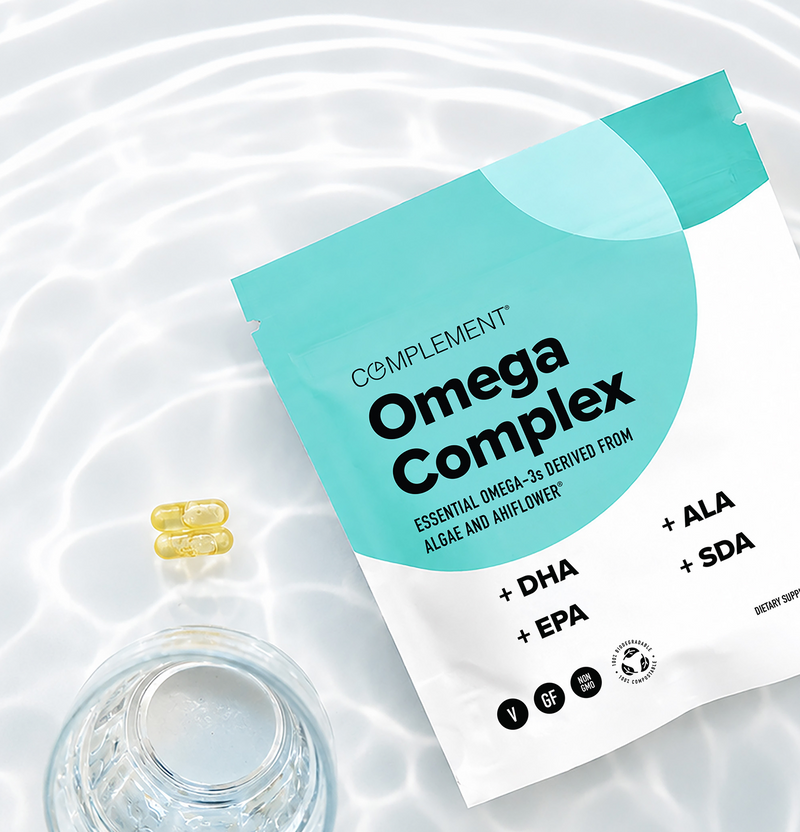 A packet labeled “Omega Complex” lies on a white surface next to a glass of water and two yellow capsules, with rippled water reflections in the background.