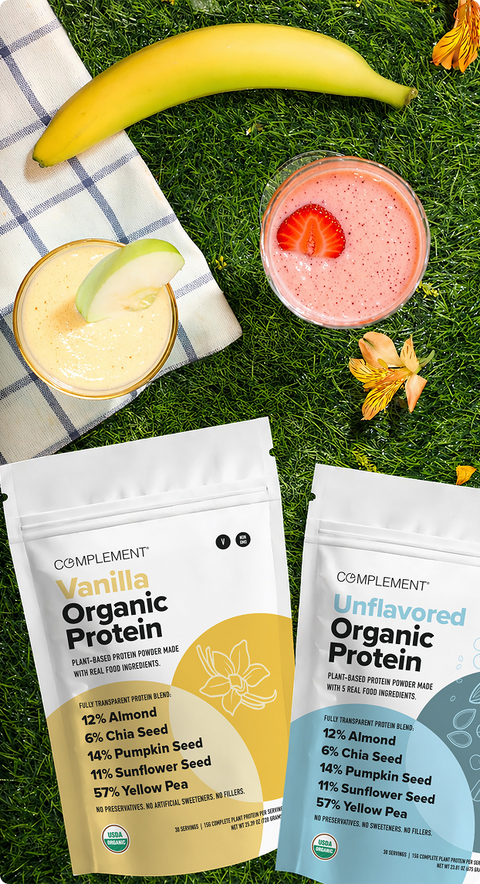 Two Complement Organic Protein powder pouches (Vanilla and Unflavored) on grass beside a banana, two smoothies, a striped napkin, and flowers. Ingredients and protein content are visible on the pouches.