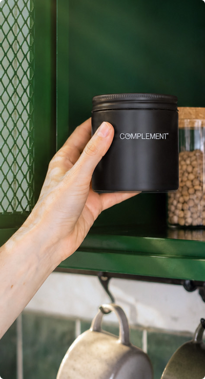 A hand places a black container labeled COMPLEMENT on a green kitchen shelf next to a glass jar filled with legumes. Metal pots hang below the shelf.