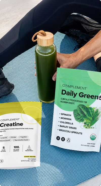 A person sits on a blue exercise mat holding a green smoothie in a glass bottle. Nearby are two supplement packets labeled “Complement Daily Greens” and “Creatine.” The person is wearing black athletic clothing and sneakers.