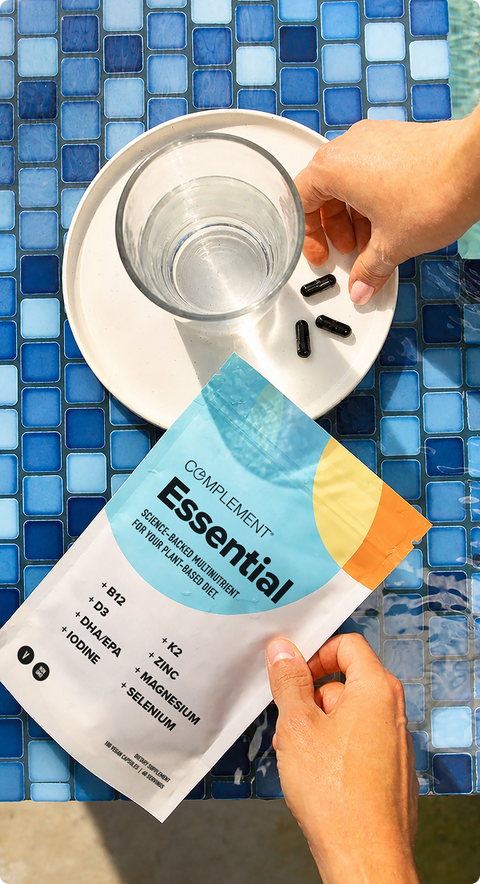 A person holds a packet of Complement Essential supplements by a plate with a glass of water and three black capsules on a blue tiled surface.