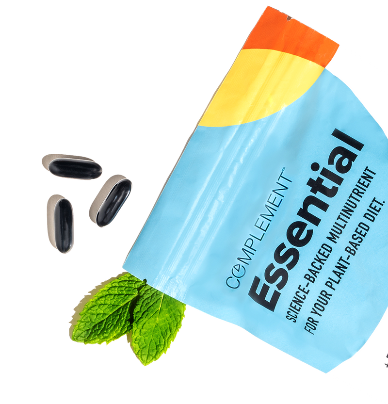 A blue and white Complement Essential supplement pouch, partially open, with three black capsules and sprigs of fresh mint arranged next to it on a white background.